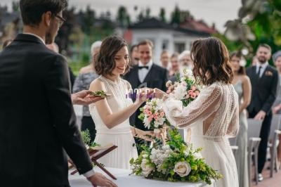 Deux femmes en robes blanches échangent des alliances lors d'une cérémonie de mariage en extérieur, entourées d'invités.