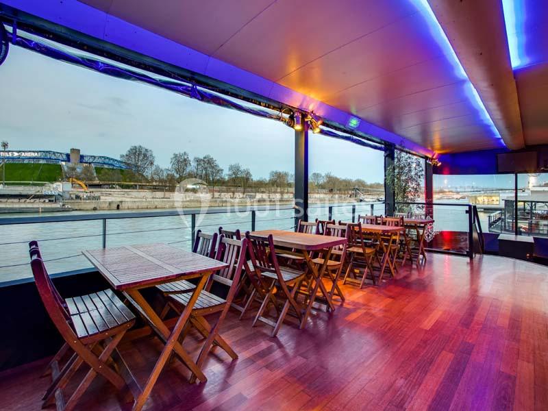 Terrasse couverte avec tables et chaises en bois, offrant une vue sur une rivière et des arbres en arrière-plan.