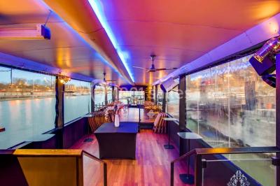 Salle avec tables et chaises en bois sur une péniche, offrant une vue sur un pont et une rivière en arrière-plan.