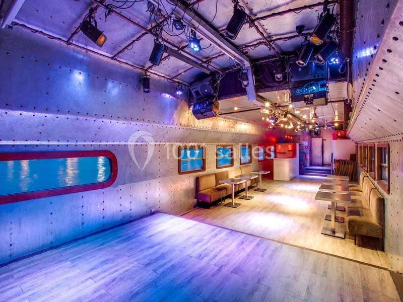 Salle au design inspiré d'un sous-marin, avec hublots, éclairage coloré et tables alignées le long des murs.