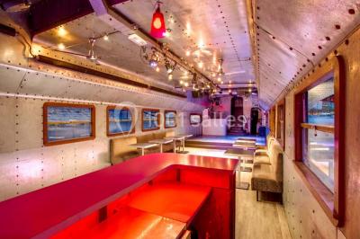 Salle avec tables et chaises en bois sur une péniche, offrant une vue sur un pont et une rivière en arrière-plan.