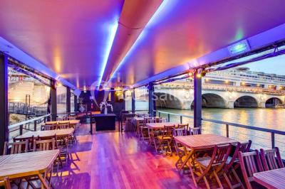 Salle avec tables et chaises en bois sur une péniche, offrant une vue sur un pont et une rivière en arrière-plan.