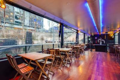 Salle avec tables et chaises en bois sur une péniche, offrant une vue sur un pont et une rivière en arrière-plan.