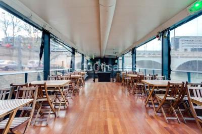 Salle avec tables et chaises en bois sur une péniche, offrant une vue sur un pont et une rivière en arrière-plan.