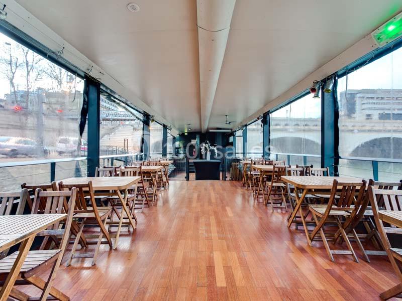 Salle avec parquet en bois, tables et chaises pliantes, baies vitrées donnant sur un paysage urbain avec pont.