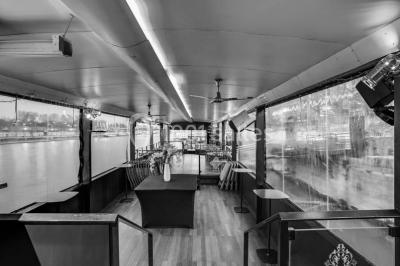 Salle avec tables et chaises en bois sur une péniche, offrant une vue sur un pont et une rivière en arrière-plan.