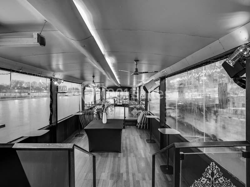 Intérieur d'une péniche avec parquet, tables, chaises et grandes baies vitrées donnant sur un paysage fluvial.