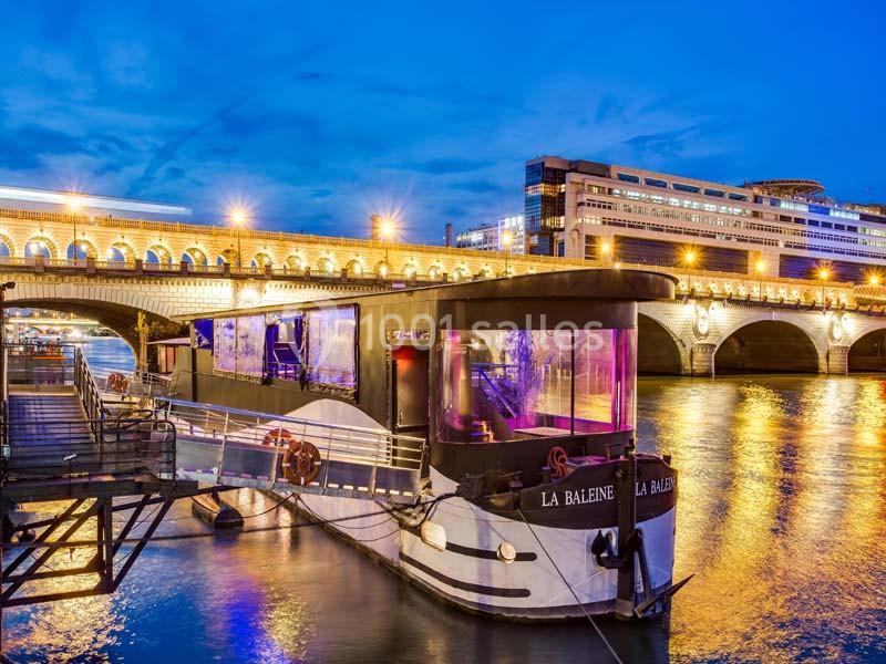 Bateau amarré sur une rivière illuminée, avec un pont et des bâtiments en arrière-plan au crépuscule.