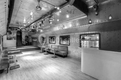 Salle avec tables et chaises en bois sur une péniche, offrant une vue sur un pont et une rivière en arrière-plan.