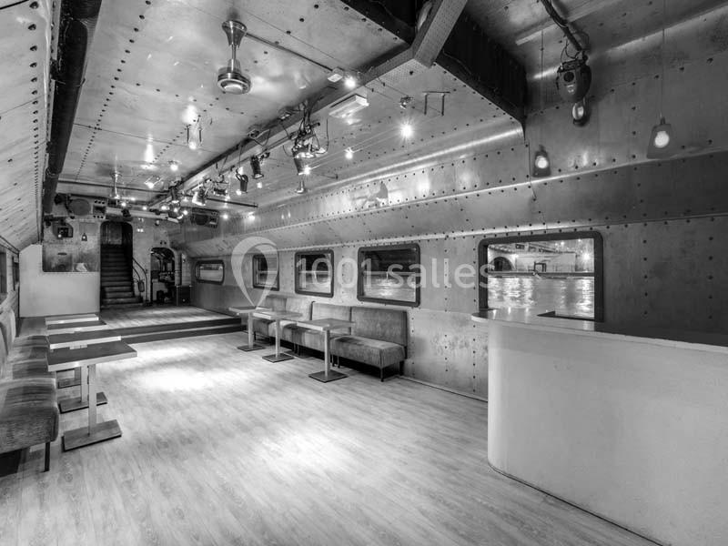 Intérieur d'une salle moderne avec murs métalliques, banquettes, tables et un comptoir, éclairée par des spots.