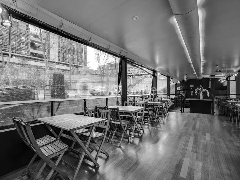 Salle de restaurant vide avec des tables et chaises en bois, grandes baies vitrées donnant sur un extérieur urbain.