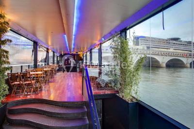 Salle avec tables et chaises en bois sur une péniche, offrant une vue sur un pont et une rivière en arrière-plan.
