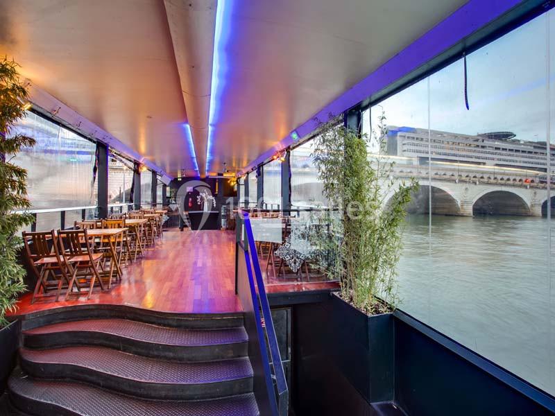 Intérieur d'une péniche aménagée avec des tables et des chaises, vue sur un pont et une rivière à travers des baies vitrées.