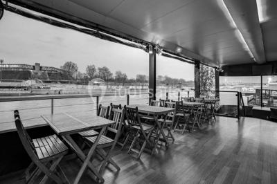 Salle avec tables et chaises en bois sur une péniche, offrant une vue sur un pont et une rivière en arrière-plan.