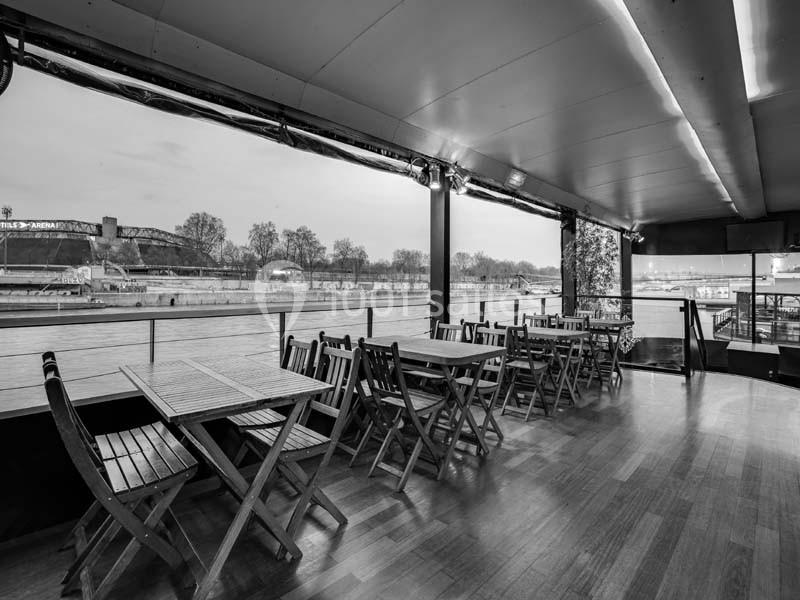 Terrasse couverte avec tables et chaises en bois, offrant une vue sur un fleuve et des arbres en arrière-plan.