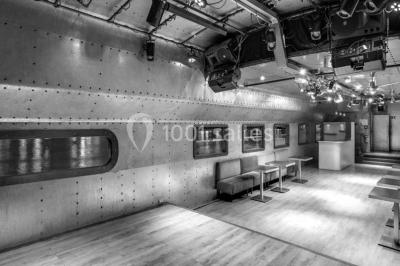 Salle avec tables et chaises en bois sur une péniche, offrant une vue sur un pont et une rivière en arrière-plan.