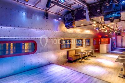 Salle avec tables et chaises en bois sur une péniche, offrant une vue sur un pont et une rivière en arrière-plan.