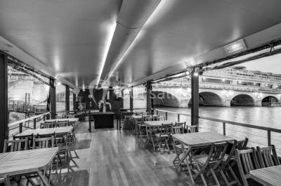 Salle avec tables et chaises en bois sur une péniche, offrant une vue sur un pont et une rivière en arrière-plan.