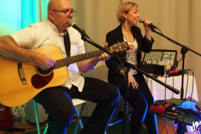 Un duo musical se produit sur une scène intérieure avec guitare et chant, devant un public assis.