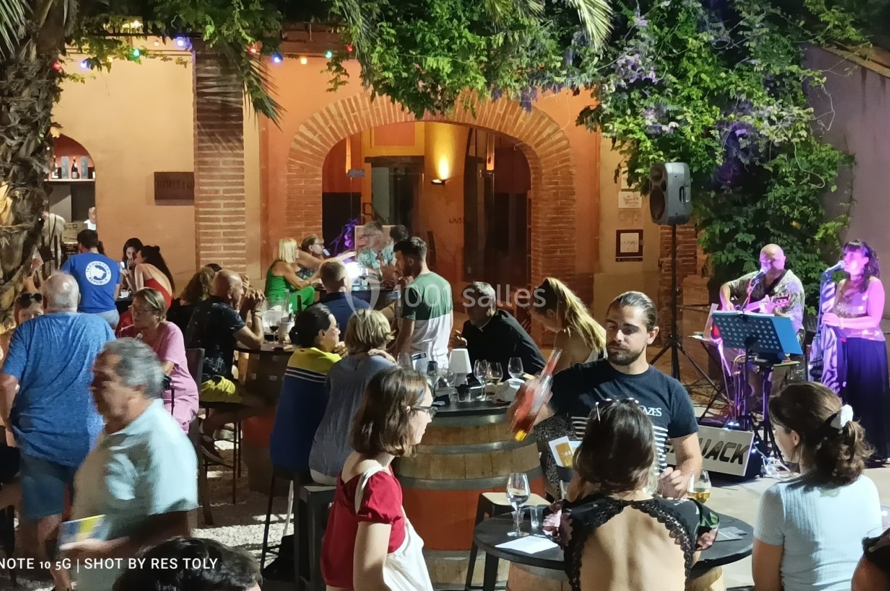 Groupe de personnes attablées en soirée dans une cour extérieure éclairée, avec des musiciens jouant en arrière-plan.