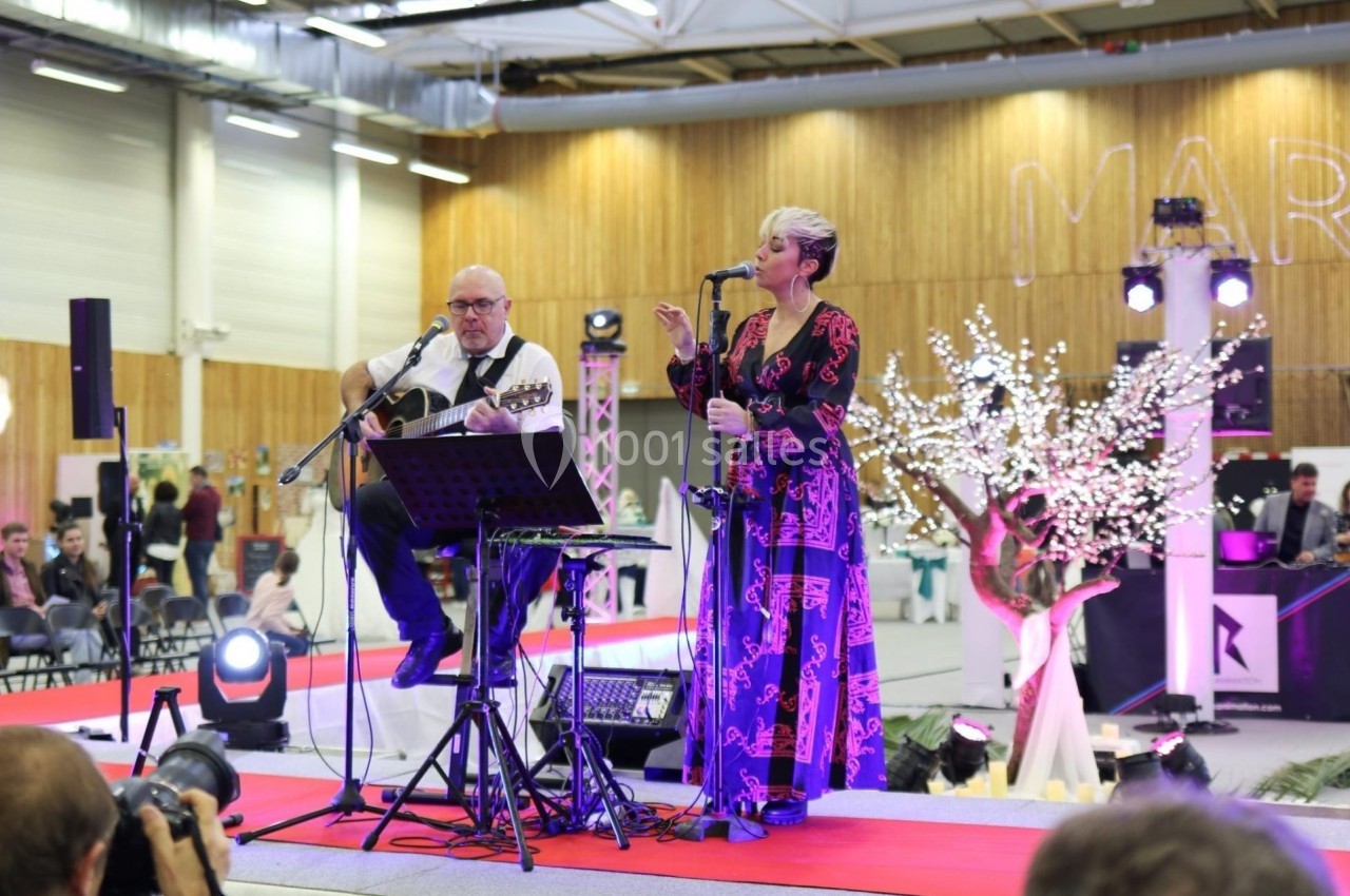 Un duo musical se produit sur une scène intérieure avec guitare et chant, devant un public assis.