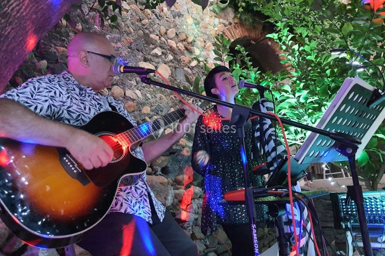 Deux musiciens jouent de la guitare et chantent en extérieur, entourés de végétation et éclairés par des lumières colorées.