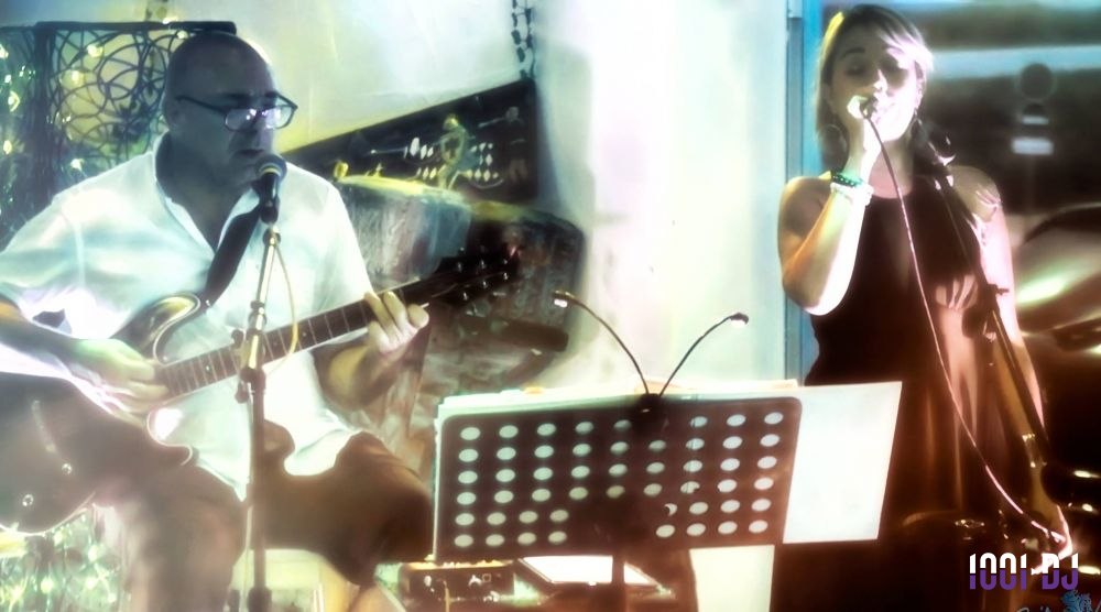 Un homme joue de la guitare et une femme chante sur une scène éclairée, avec un pupitre au premier plan.