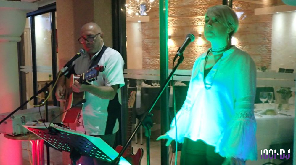 Un homme joue de la guitare et une femme chante sur scène dans un espace intérieur éclairé.