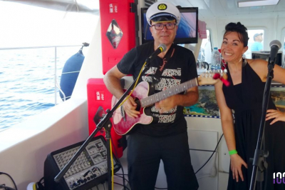 Un homme jouant de la guitare et une femme souriante posent devant des micros à bord d'un bateau.