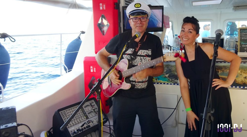Un homme jouant de la guitare et une femme souriante posent devant des micros à bord d'un bateau.