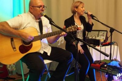 Un homme joue de la guitare acoustique et une femme chante, tous deux assis devant des micros dans un cadre lumineux.