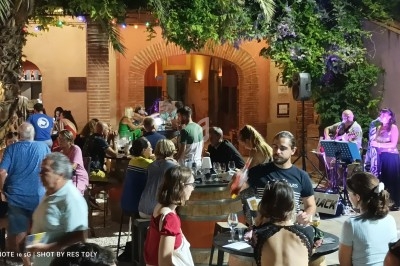 Groupe de personnes attablées en soirée dans une cour extérieure éclairée, avec des musiciens jouant en arrière-plan.