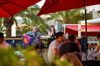 Un homme joue de la guitare et une femme chante sur une scène en plein air, entourés de parasols rouges et de palmiers.