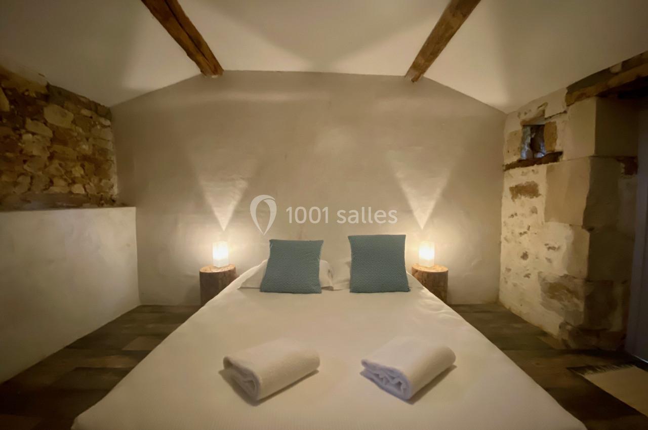 Lit double avec draps blancs, deux coussins bleus, éclairage doux et murs en pierre dans une chambre rustique.