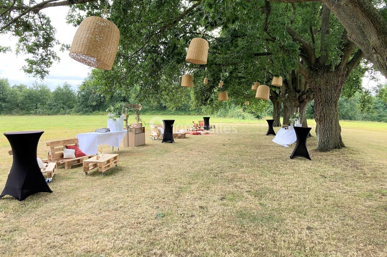 Espace extérieur aménagé sous des arbres avec des tables hautes, des palettes en bois et des lampes suspendues.