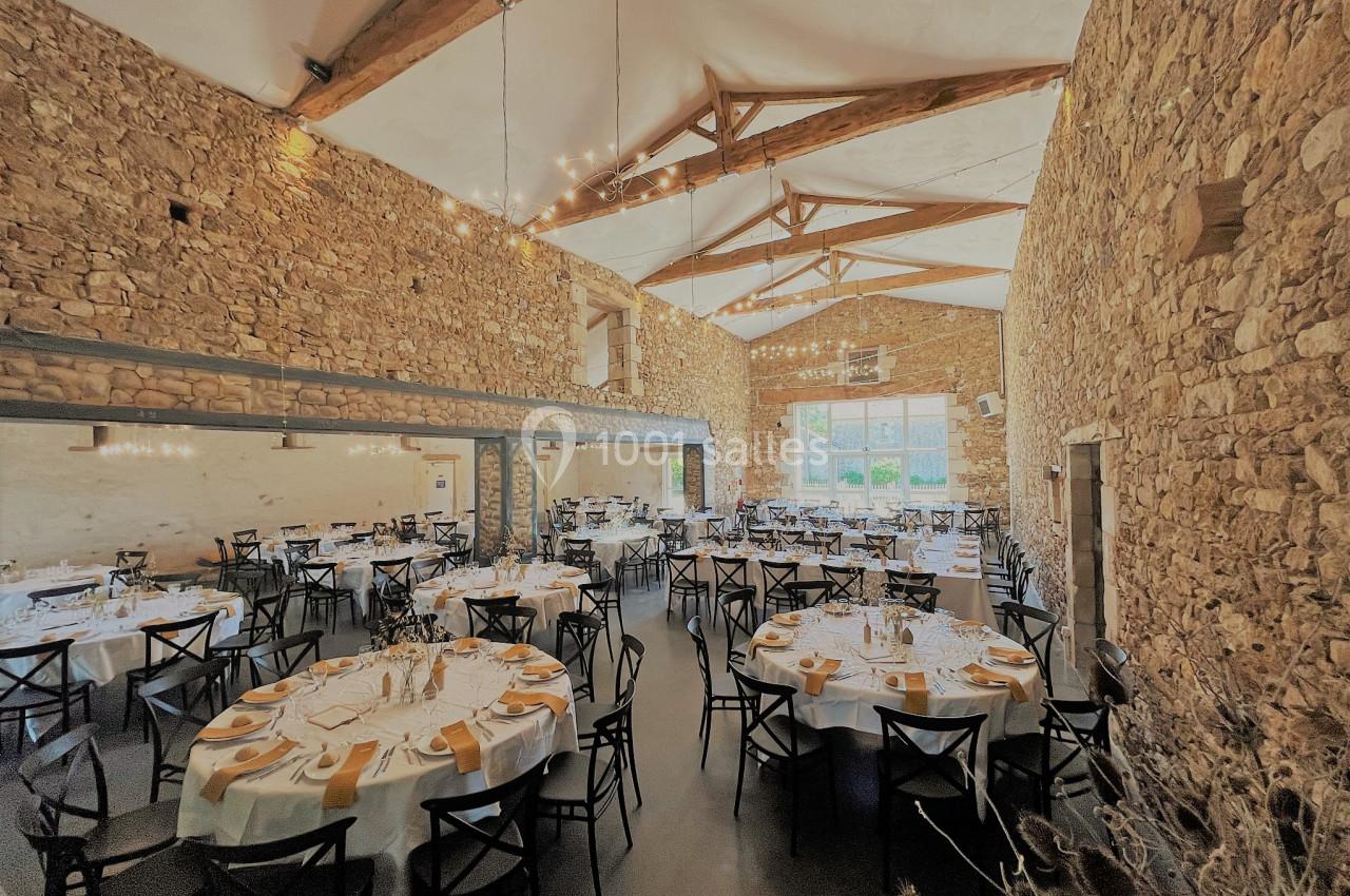 Salle de réception en pierre avec tables rondes dressées, nappes blanches et serviettes jaunes, éclairée par des lustres.