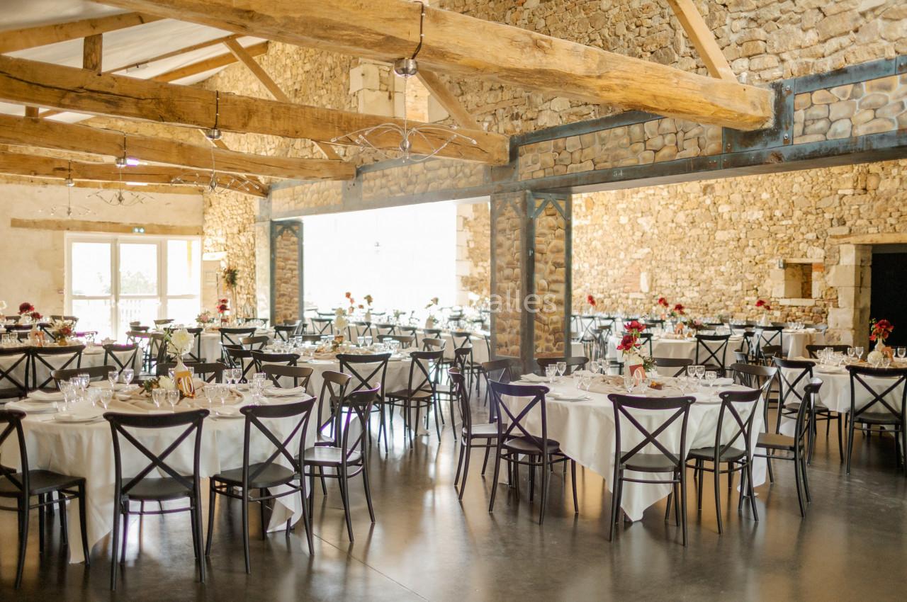 Salle de réception lumineuse avec murs en pierre, poutres apparentes et tables dressées pour un événement.