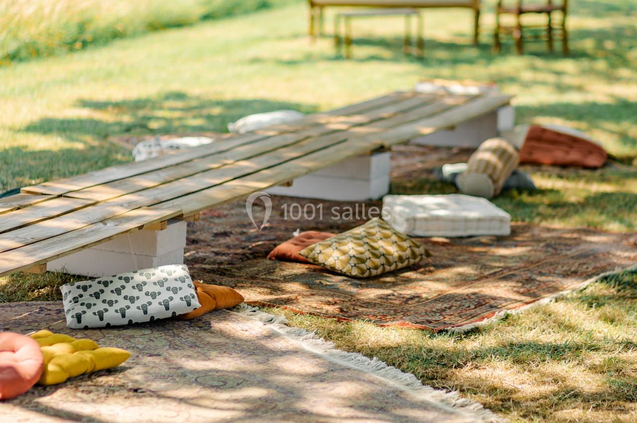 Banc en bois avec coussins colorés sur des tapis en plein air, entouré d'herbe et de mobilier vintage.