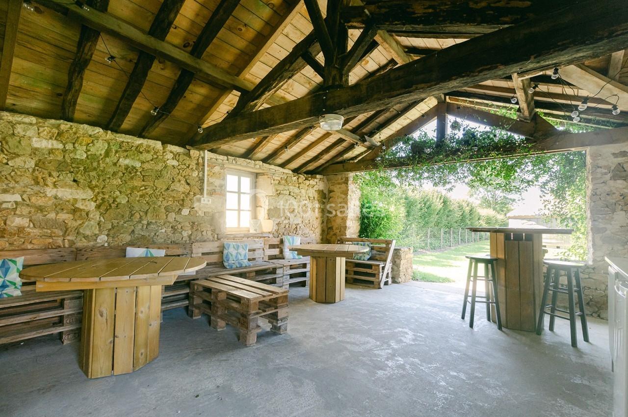 Terrasse couverte en pierre et bois avec mobilier rustique, ouverte sur un jardin verdoyant.