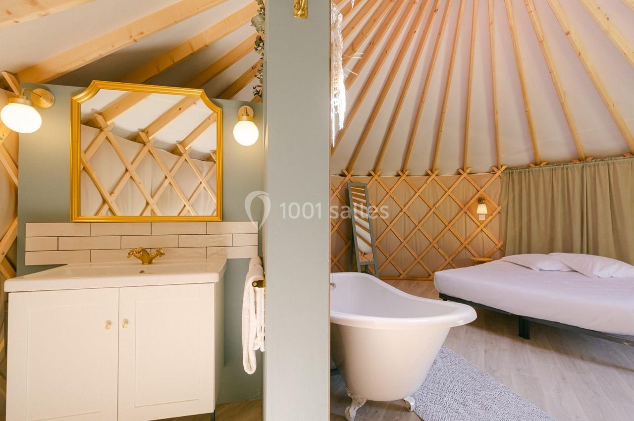 Intérieur d'une yourte avec salle de bain équipée d'une baignoire, lavabo et miroir doré, et un lit en arrière-plan.