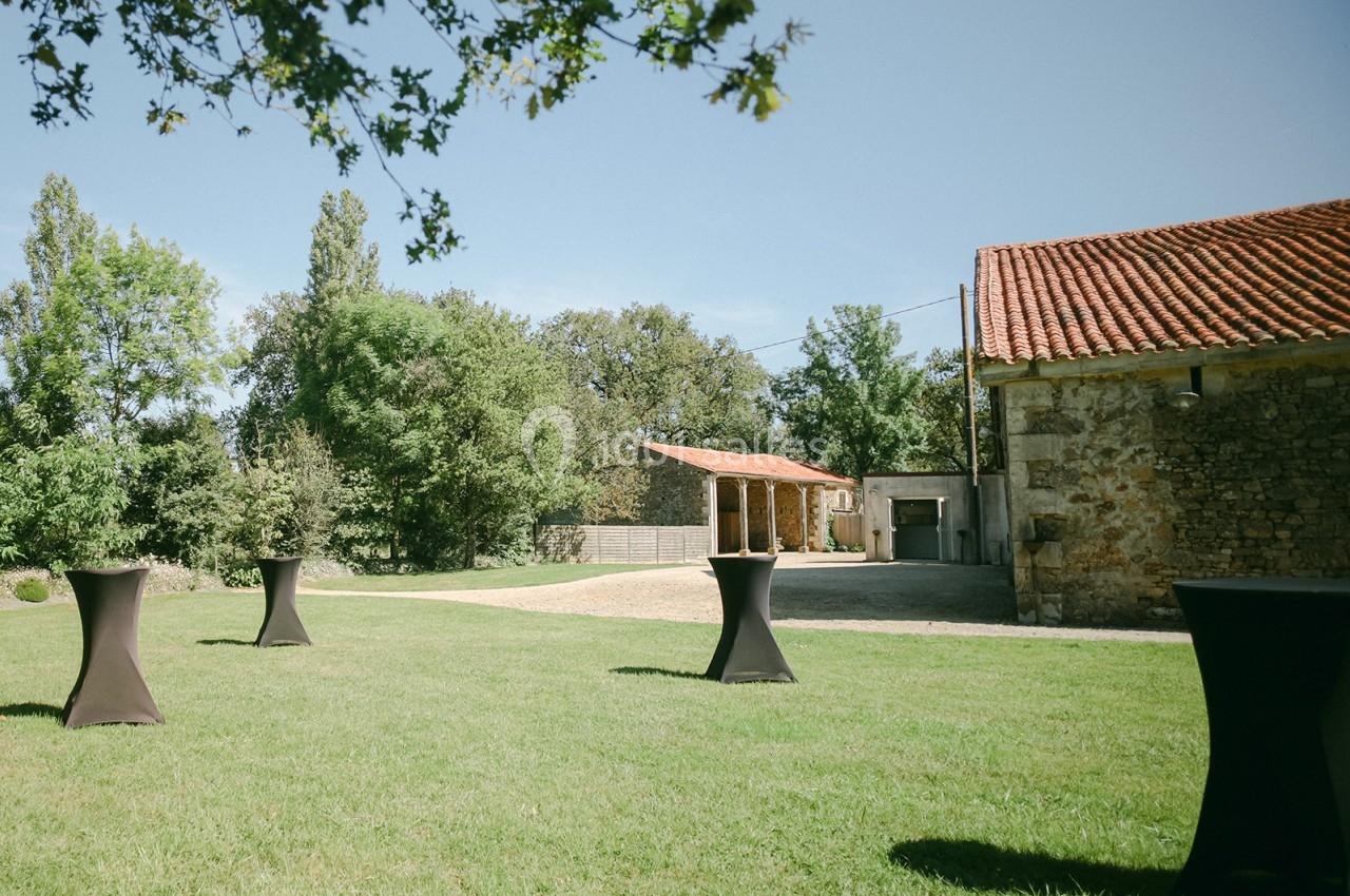 Cour extérieure avec pelouse, tables hautes noires, bâtiments en pierre et toit en tuiles sous un ciel dégagé.