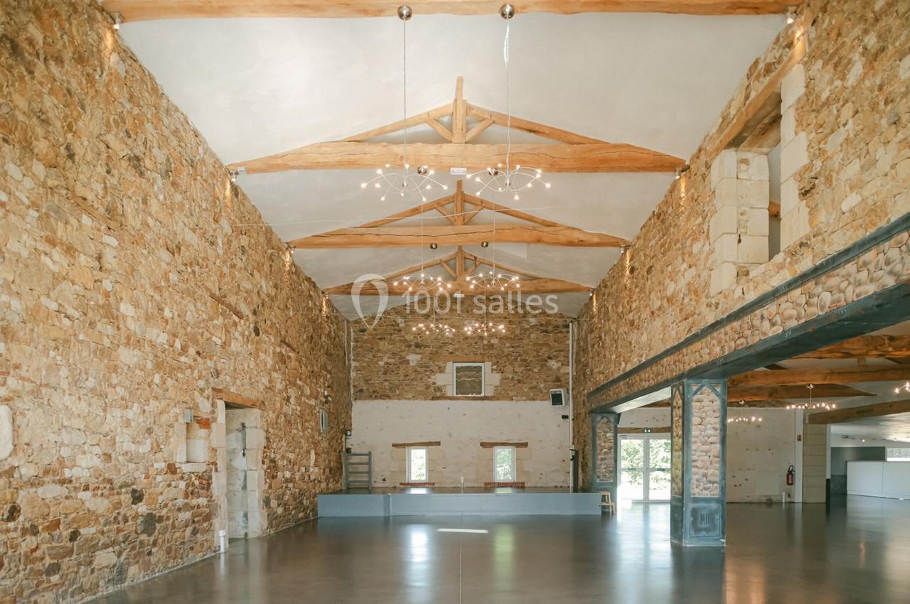 Salle spacieuse avec murs en pierre, poutres apparentes et éclairage suspendu, offrant une ambiance lumineuse et rustique.