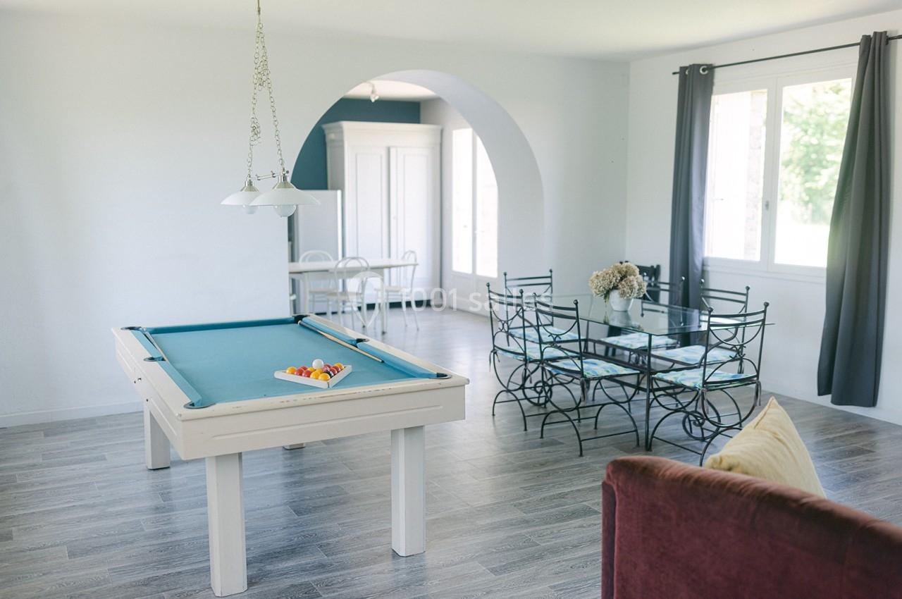 Salle lumineuse avec table de billard, coin repas avec chaises en métal et grande fenêtre donnant sur l'extérieur.