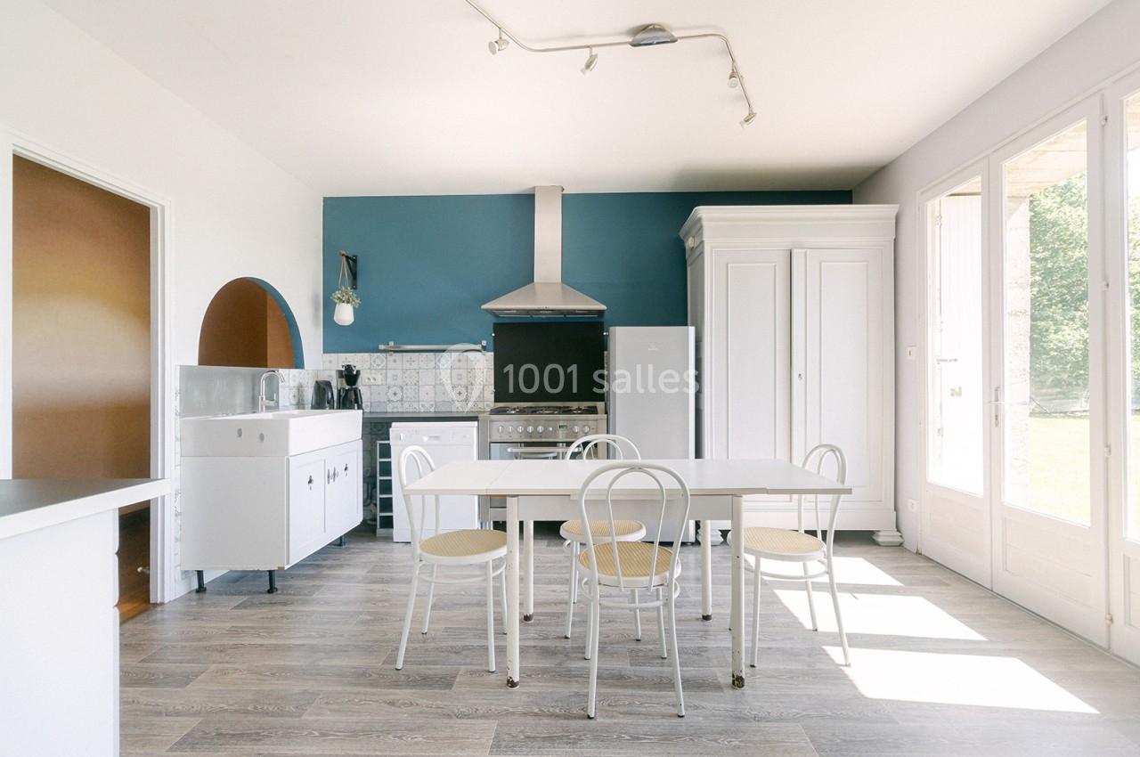 Cuisine lumineuse avec table blanche, chaises en métal, mur bleu, sol en bois clair et grande fenêtre donnant sur l…