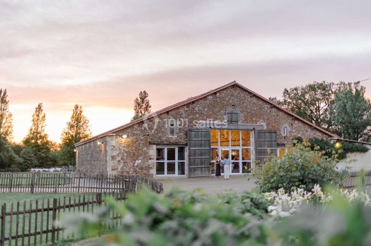 Bâtiment en pierre avec grandes portes ouvertes, entouré de verdure, au coucher du soleil.