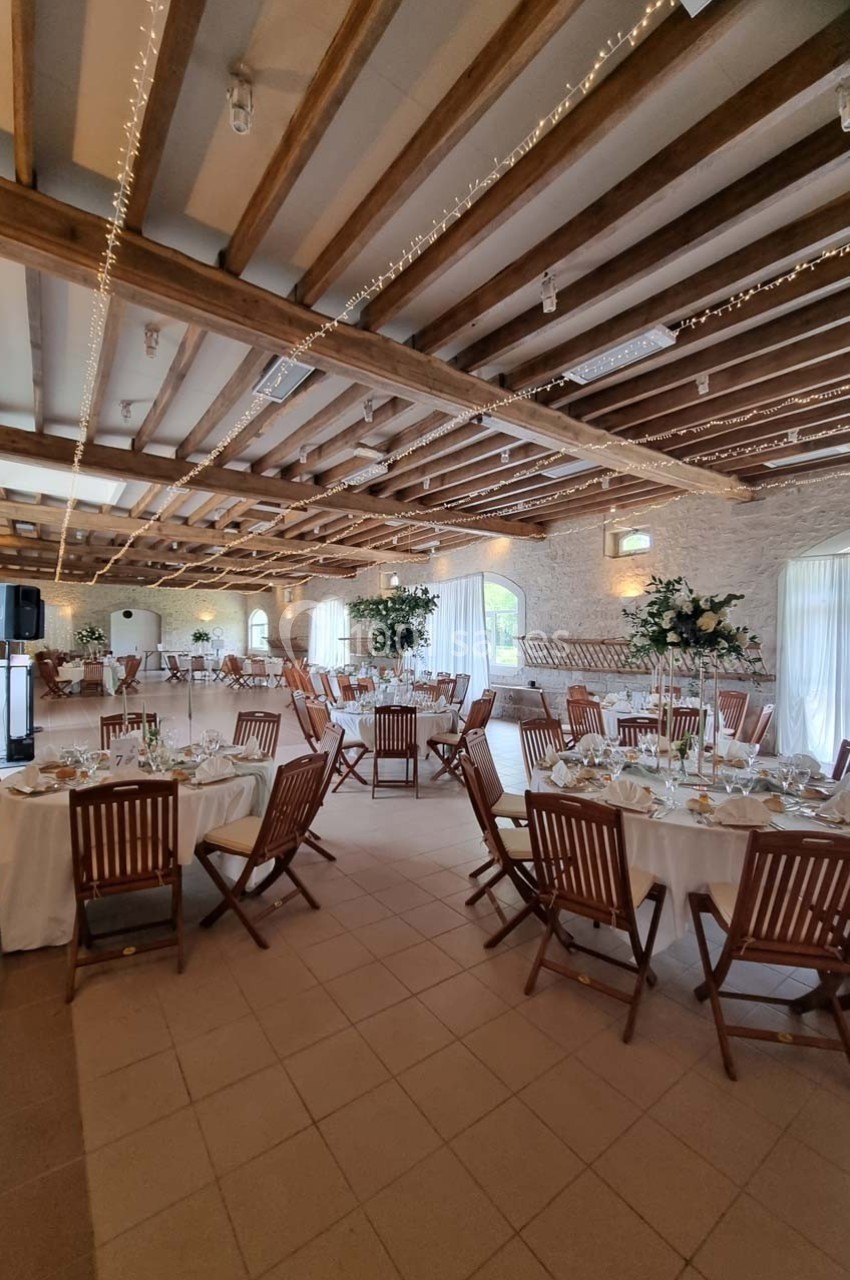 Salle de réception lumineuse avec tables rondes dressées, chaises en bois et poutres apparentes au plafond.