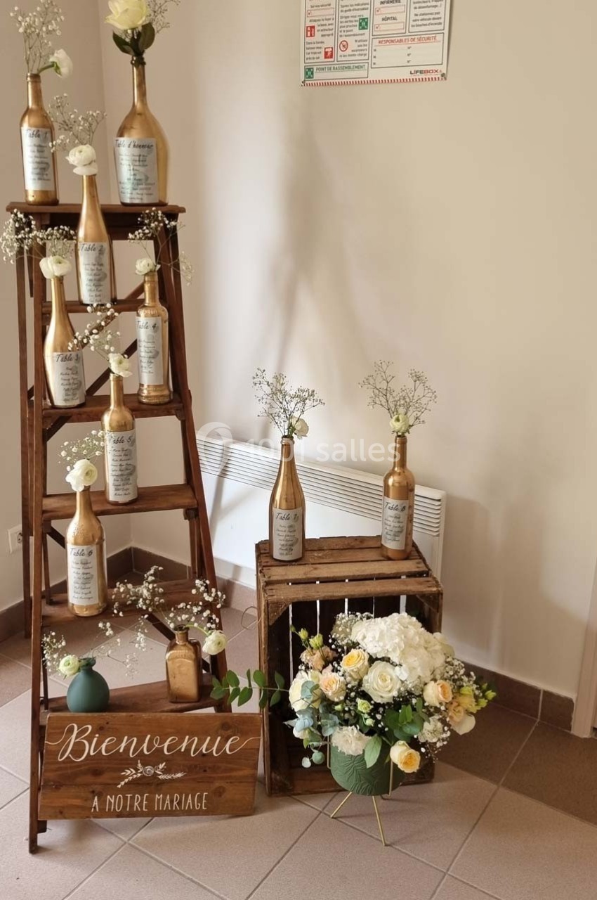 Décoration de mariage avec des bouteilles dorées sur une échelle en bois, des fleurs et un panneau ’Bienvenue à notre…