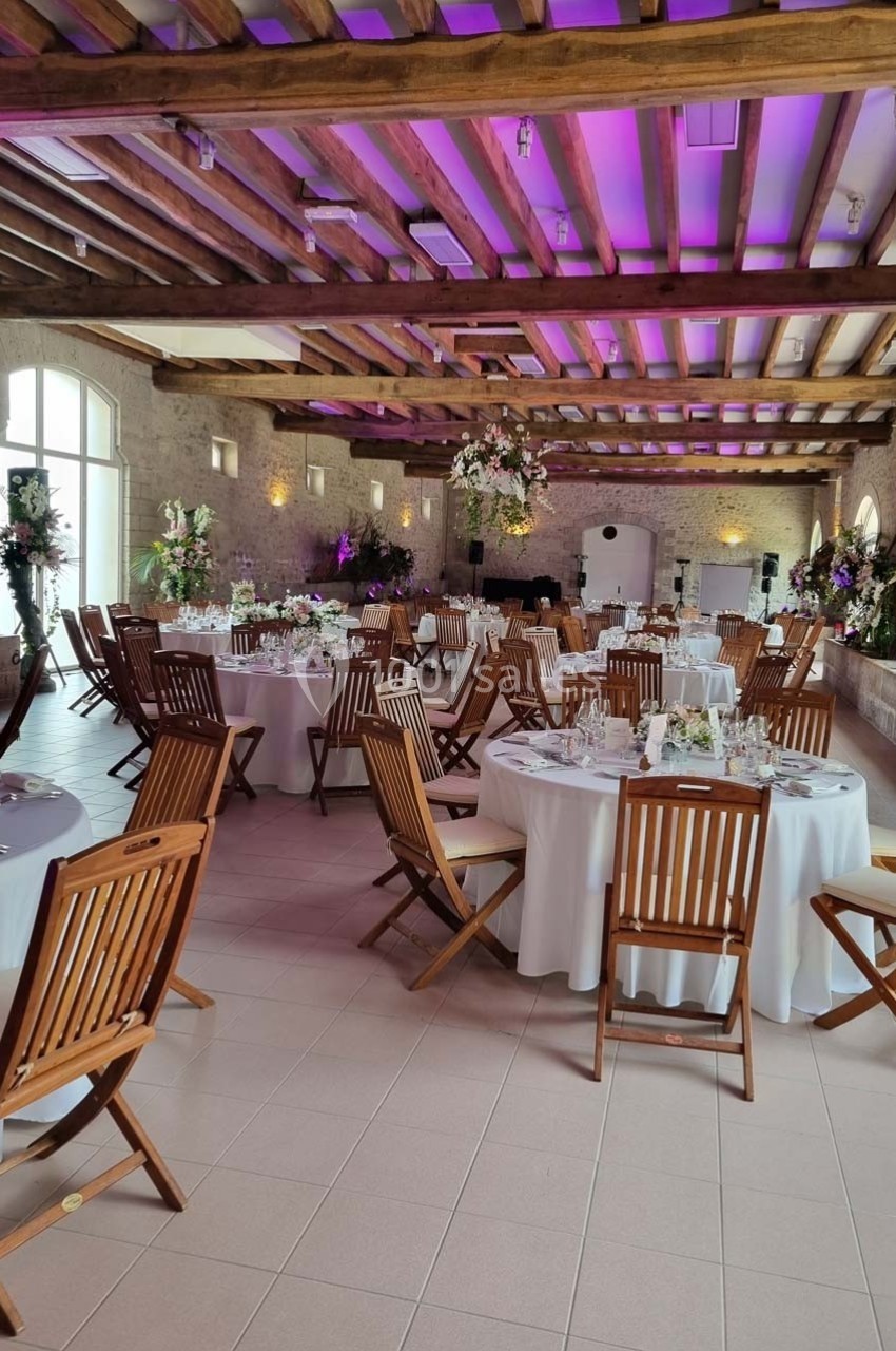 Salle de réception décorée avec des tables rondes, nappes blanches, chaises en bois et éclairage violet.