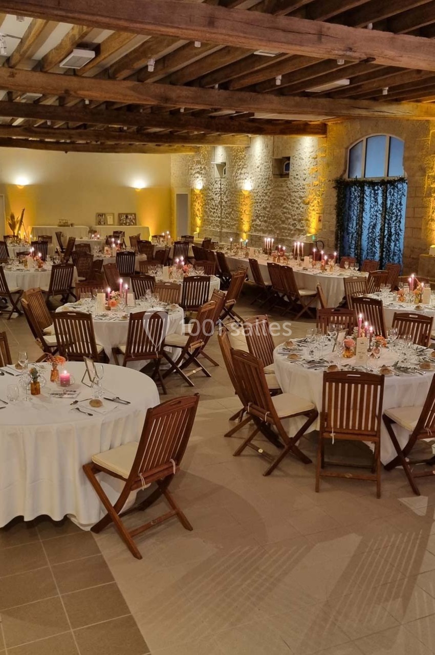 Salle de réception avec tables rondes dressées, chaises en bois, éclairage chaleureux et murs en pierre apparente.
