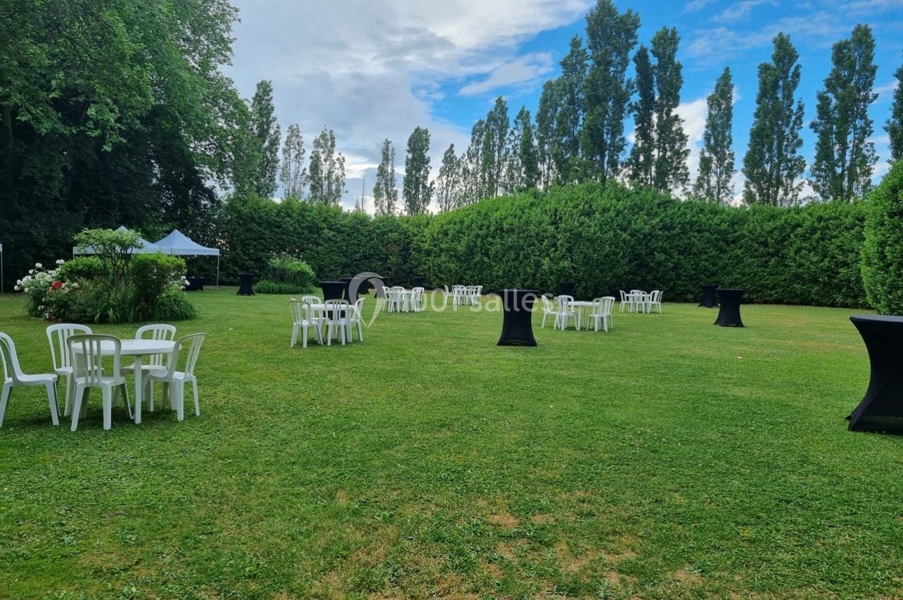 Chaises et tables blanches disposées sur une pelouse avec des mange-debout noirs, entourées de haies et d'arbres.